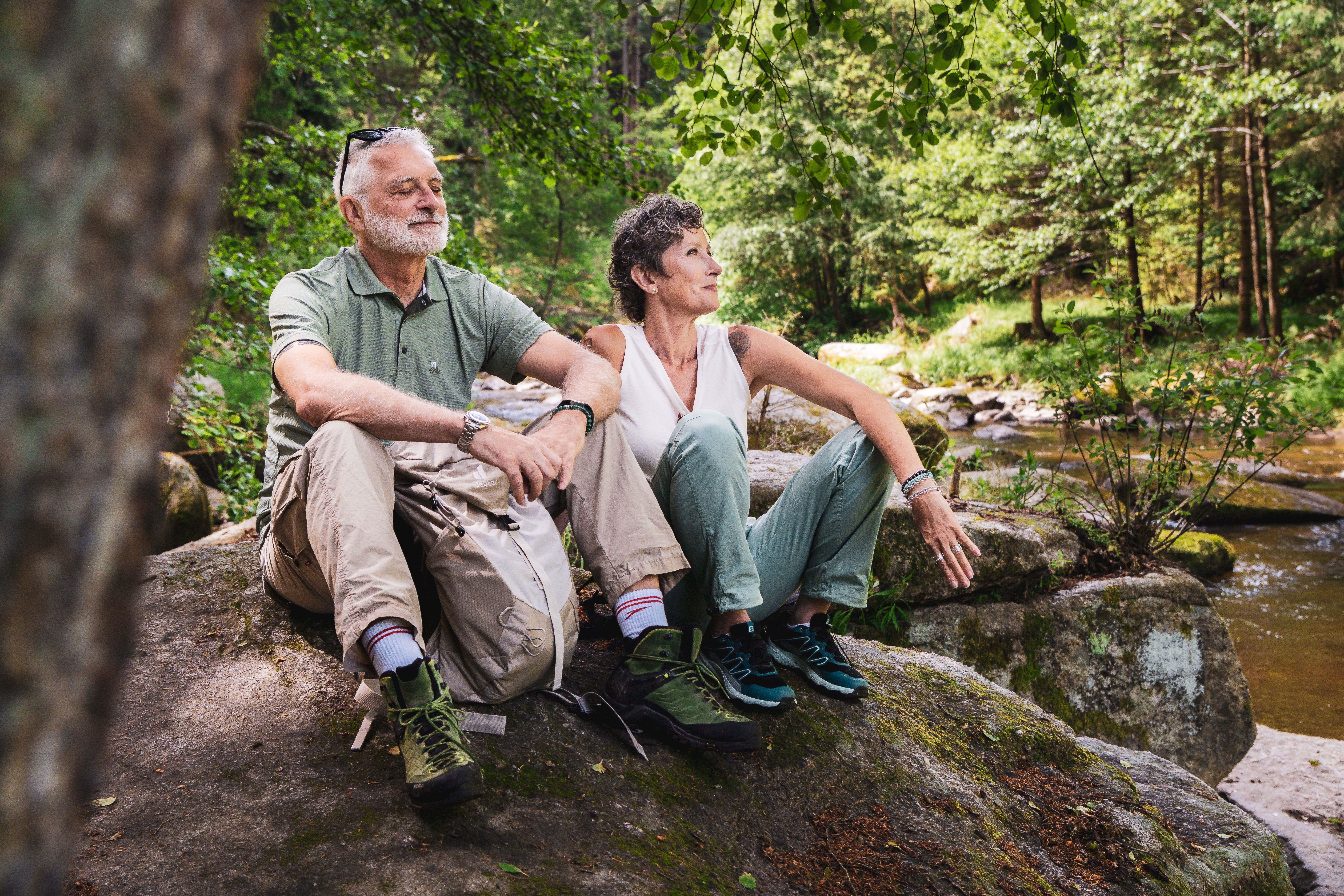 ein Mann und eine Frau mittleren Alters sitzen auf einem Stein, im Hintergrund ein Fluss