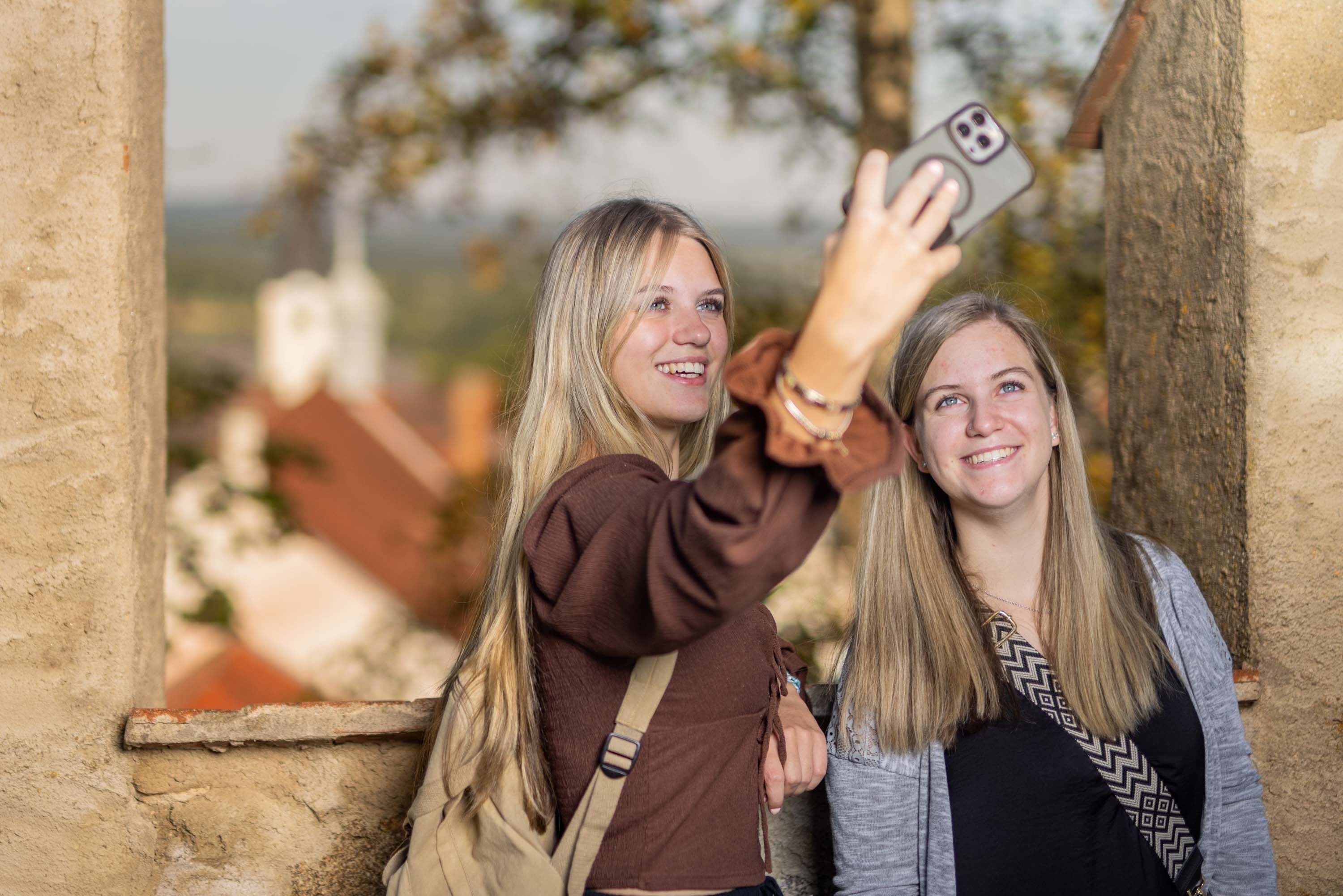 2 Frauen machen ein Selfie, links und rechts ist eine Mauer zu sehen, im Hintergrund die Stadtpfarrkirche Weitra