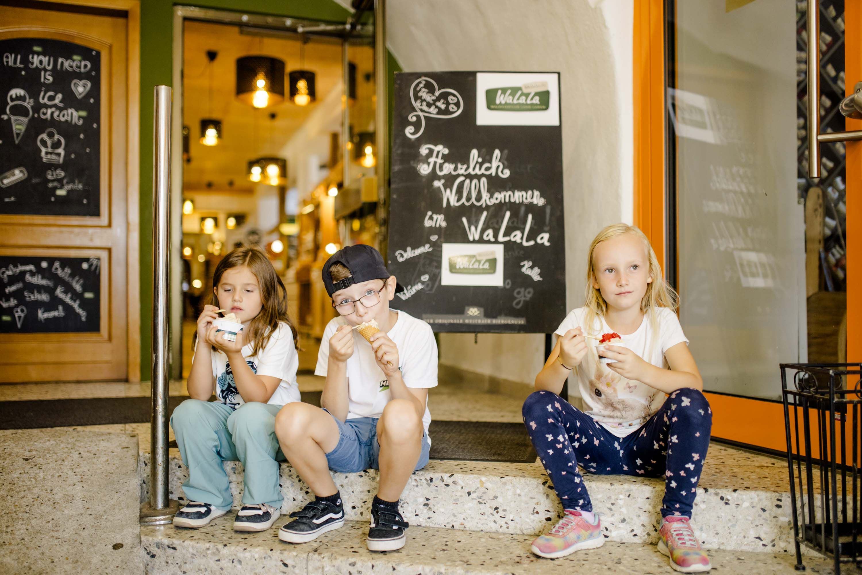 Drei Kinder beim Eis essen. Sie sitzen auf der Stufe vor einem Geschäft.