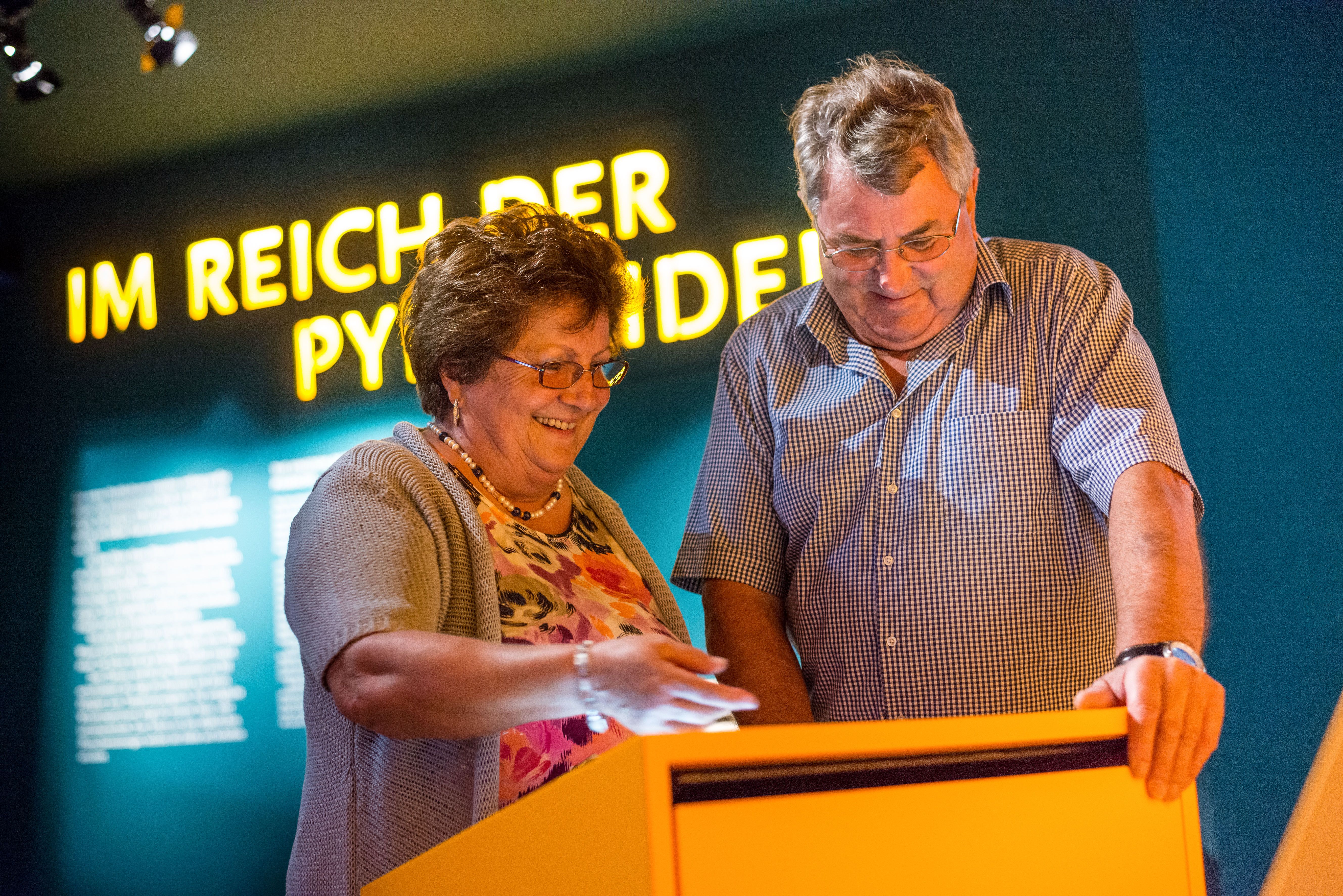 Zwei Personen betrachten ein interaktives Display in einem Museum mit der Aufschrift 'Im Reich der Pyramiden' im Hintergrund.