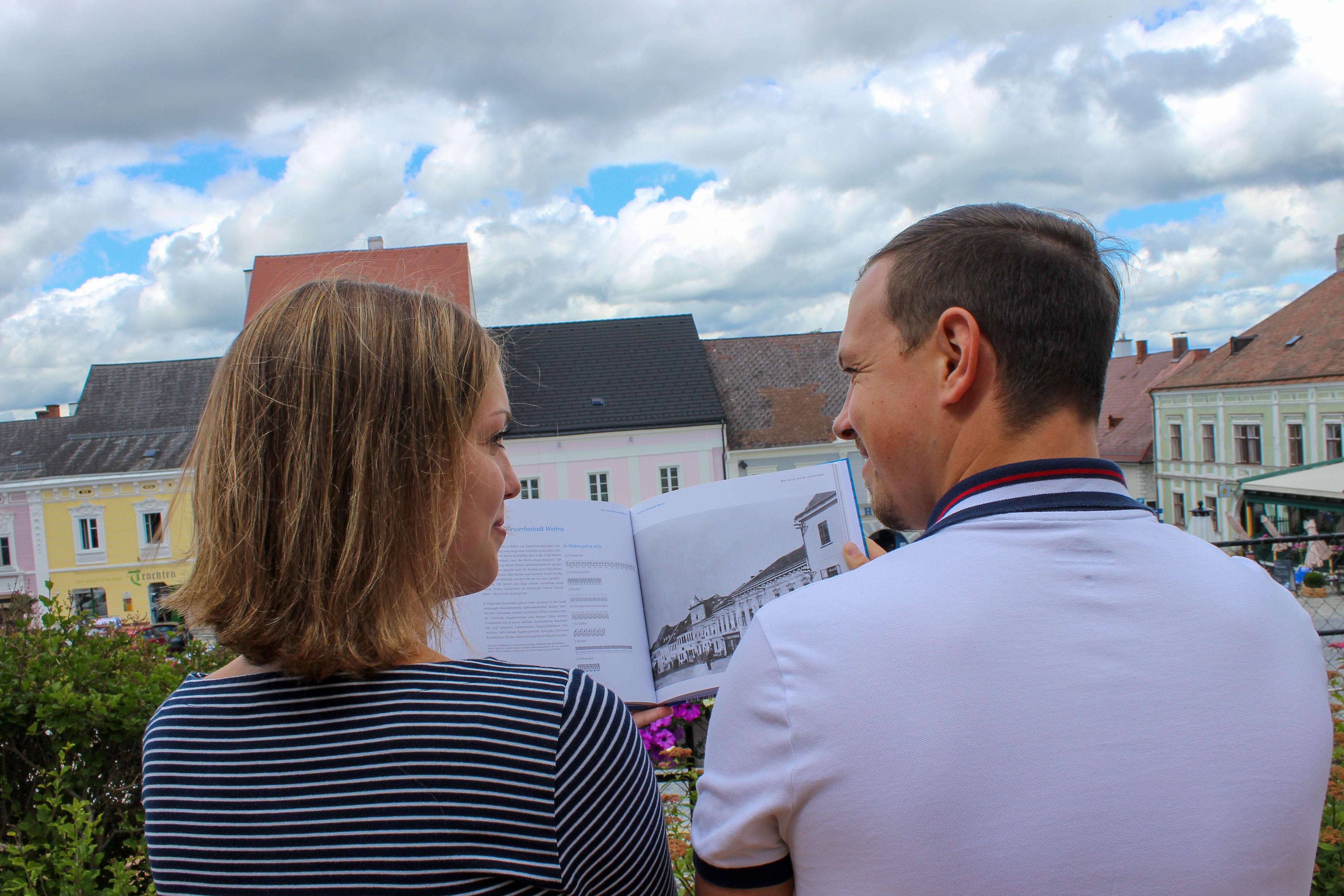 Ein Mann und eine Frau schauen sich ein Buch an. Im Hintergrund historische Bürgerhäuser in Weitra.