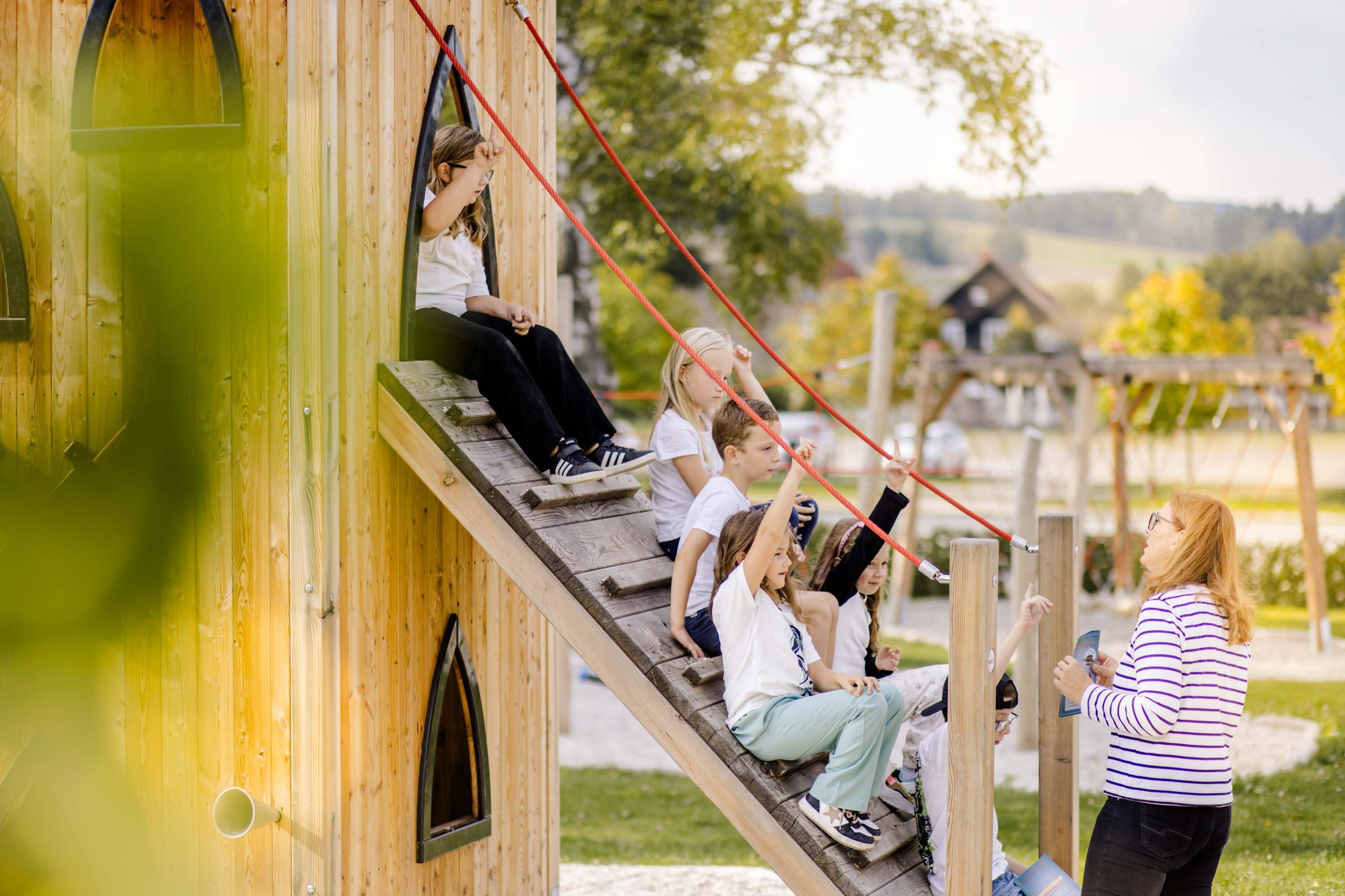 Eine Gruppe Kinder sitzt auf einer Rampe am Spielplatz, davor steht eine Lehrerin und erklärt etwas.