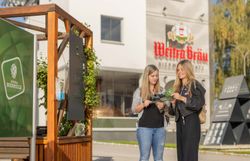 Zwei Frauen lesen eine Broschüre, links ist ein Pavillion der Weitraer Biermeile zusehen, im Hintergrund die Bierwerkstatt Weitra.