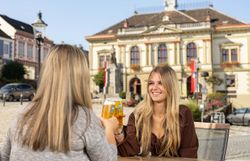 Zwei jungen Frauen sitzen an einem Tisch und prosten sich mit einem Bier zu, im Hintergrund das Rathaus von Schloss Weitra.