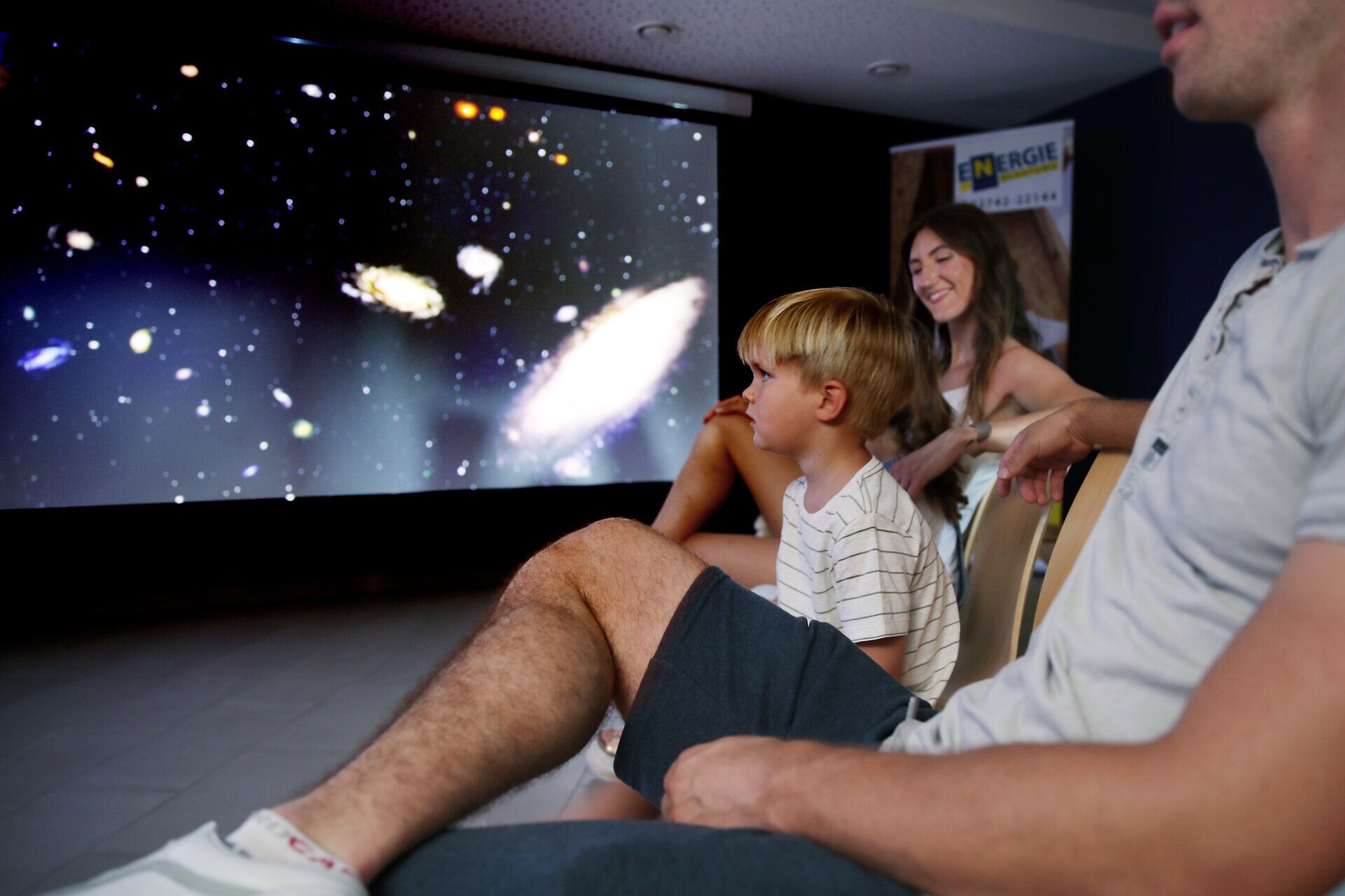 Eine Familie genießt einen spannenden Ausblick auf die unendlichen Weiten des Universums, während sie in einem gemütlichen Raum Platz genommen hat. Die funkelnden Sterne und Galaxien auf der Leinwand wecken die Neugier der kleinen Entdecker und laden zu gemeinsamen Abenteuern ein.