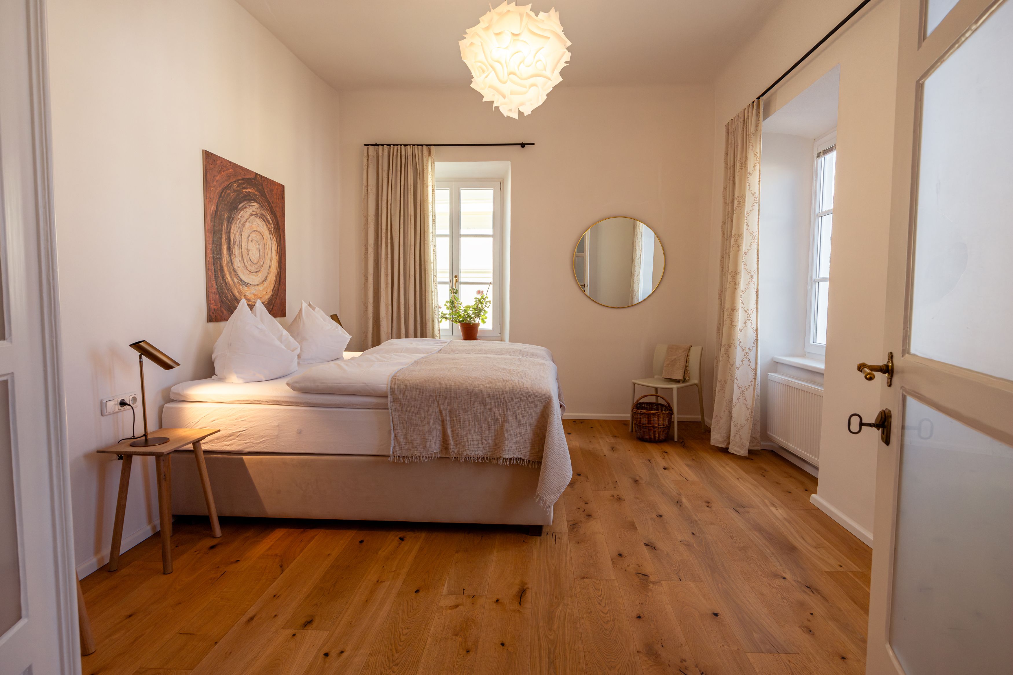 Gemütliches Schlafzimmer mit Doppelbett, Holzfußboden, Wandkunst, rundem Spiegel und Pflanzen auf dem Fensterbrett.