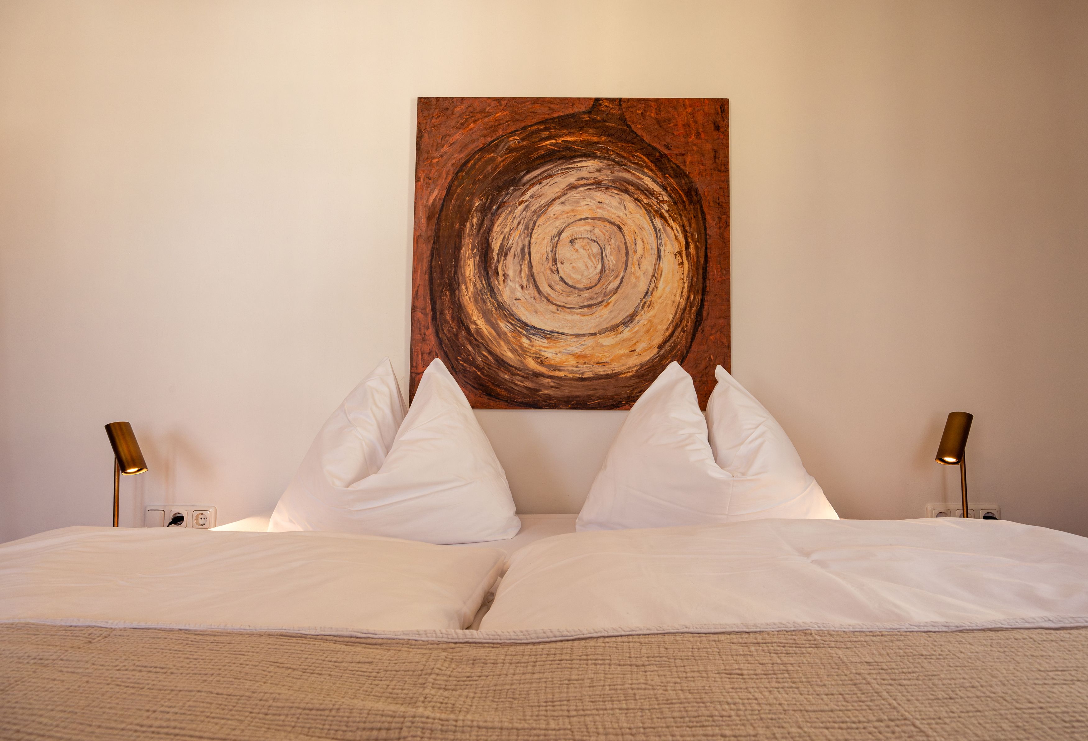 Ein gemütliches Doppelbett mit weißen Kissen und Decken, flankiert von zwei Lampen. Über dem Bett hängt ein abstraktes Gemälde mit spiralförmigem Muster.