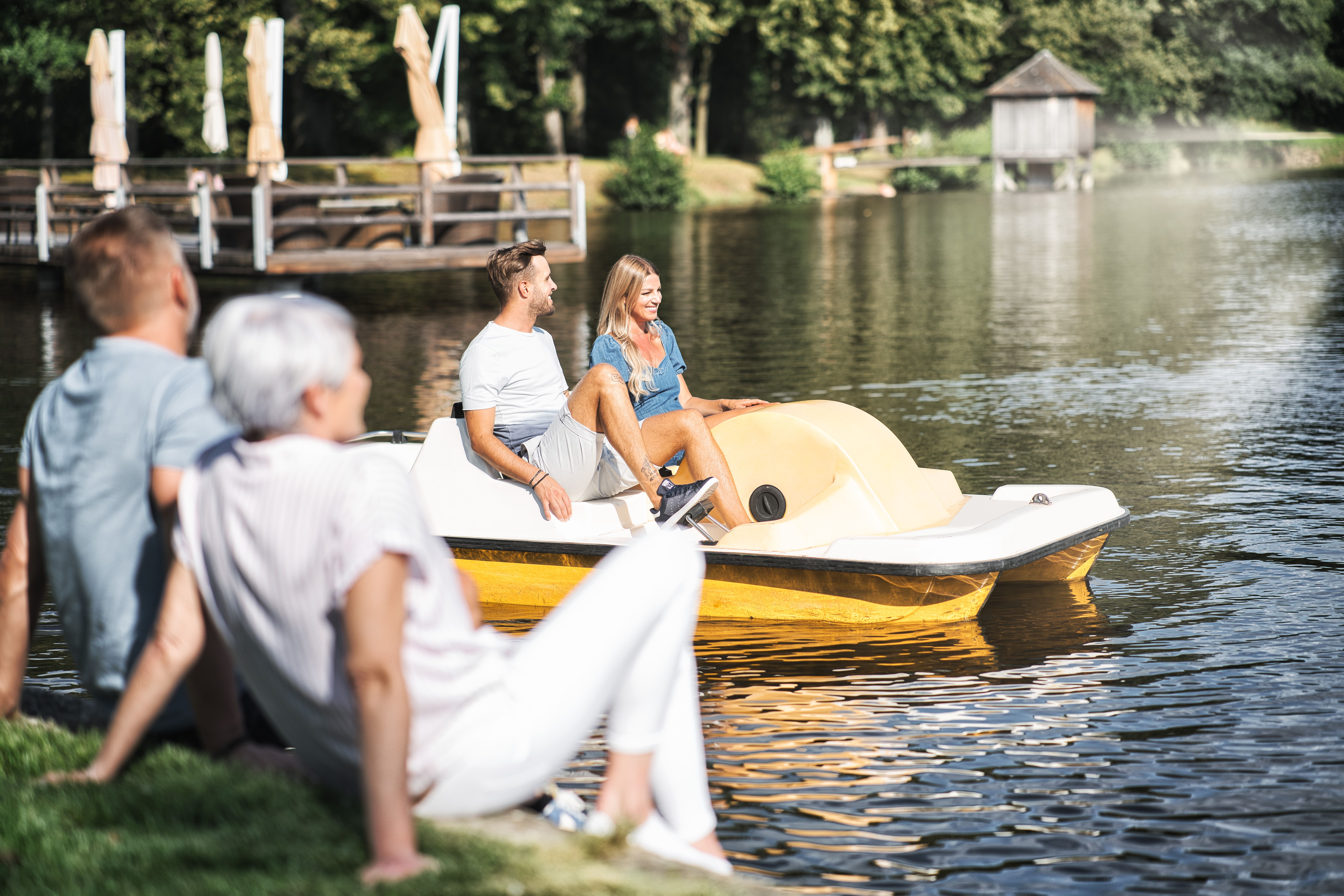 Ein Mann und eine Frau sitzen am Ufer des Hausschachenteichs, ein Mann und eine Frau fahren mit dem Trefboot vorbei.