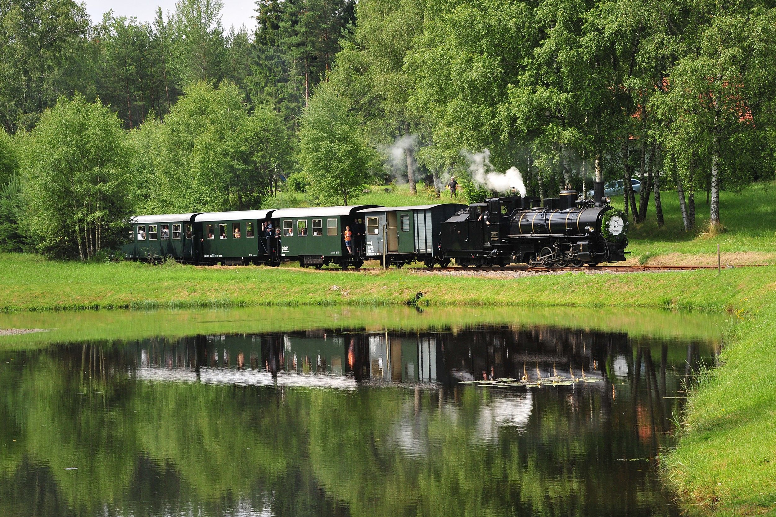 Eine historische Dampflokomotive zieht grüne Waggons durch eine grüne Landschaft, die sich in einem ruhigen Teich spiegelt.