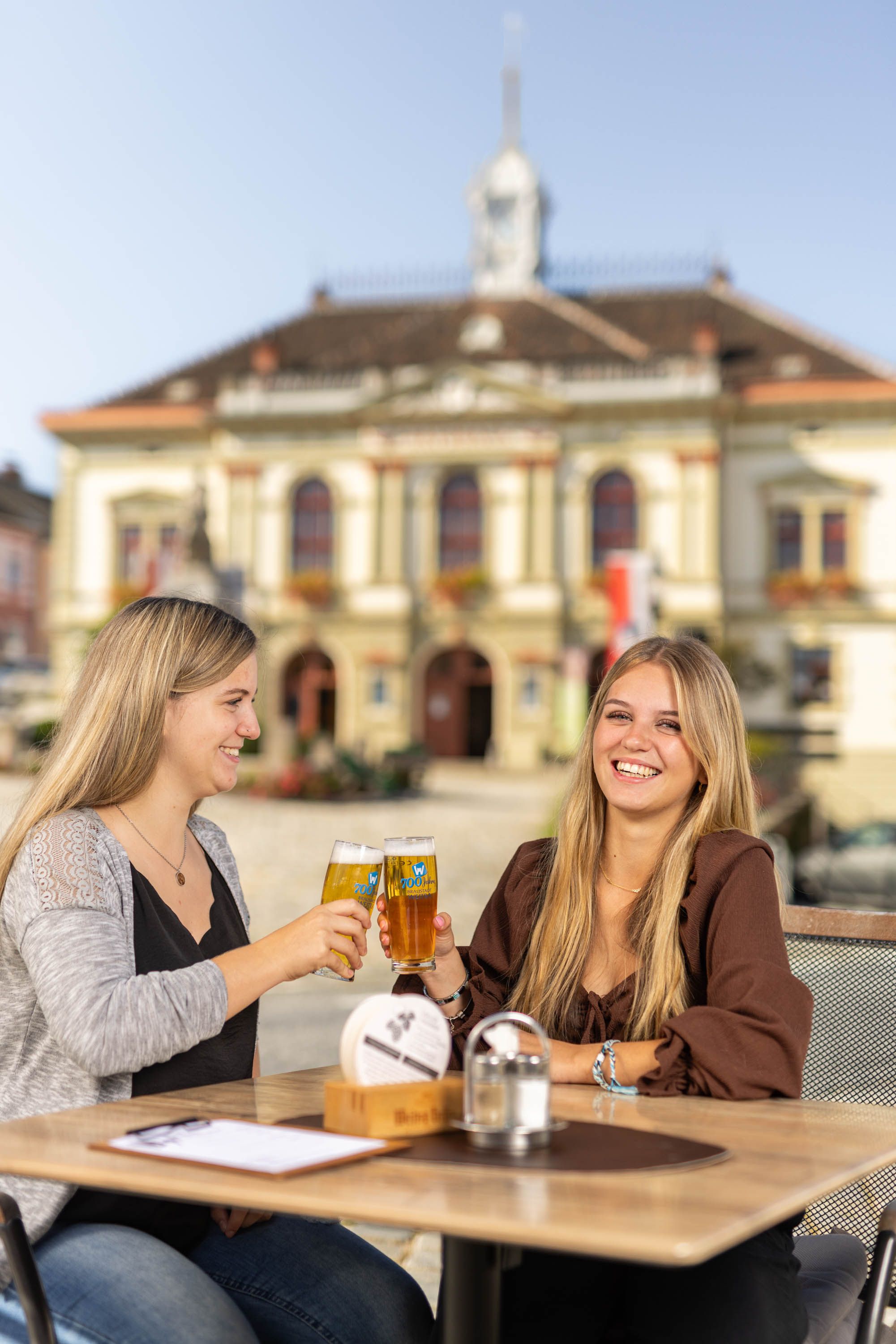 Zei Frauen prosten sich mit einem Weitraer Bier zu. Im Hintergrund das Rathaus von Weitra.