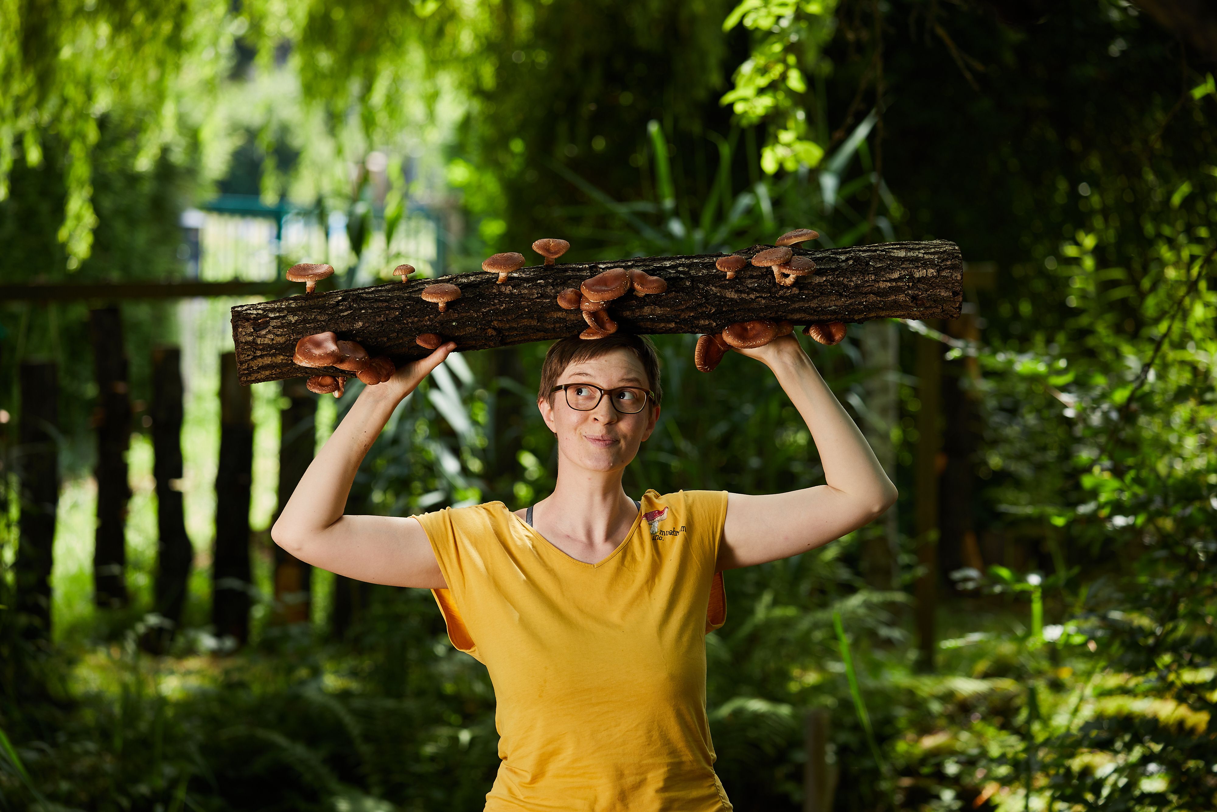 Eine Frau steht im Wald, sie hält über ihren Kopf ein Stück Totholz mit Pilzen.