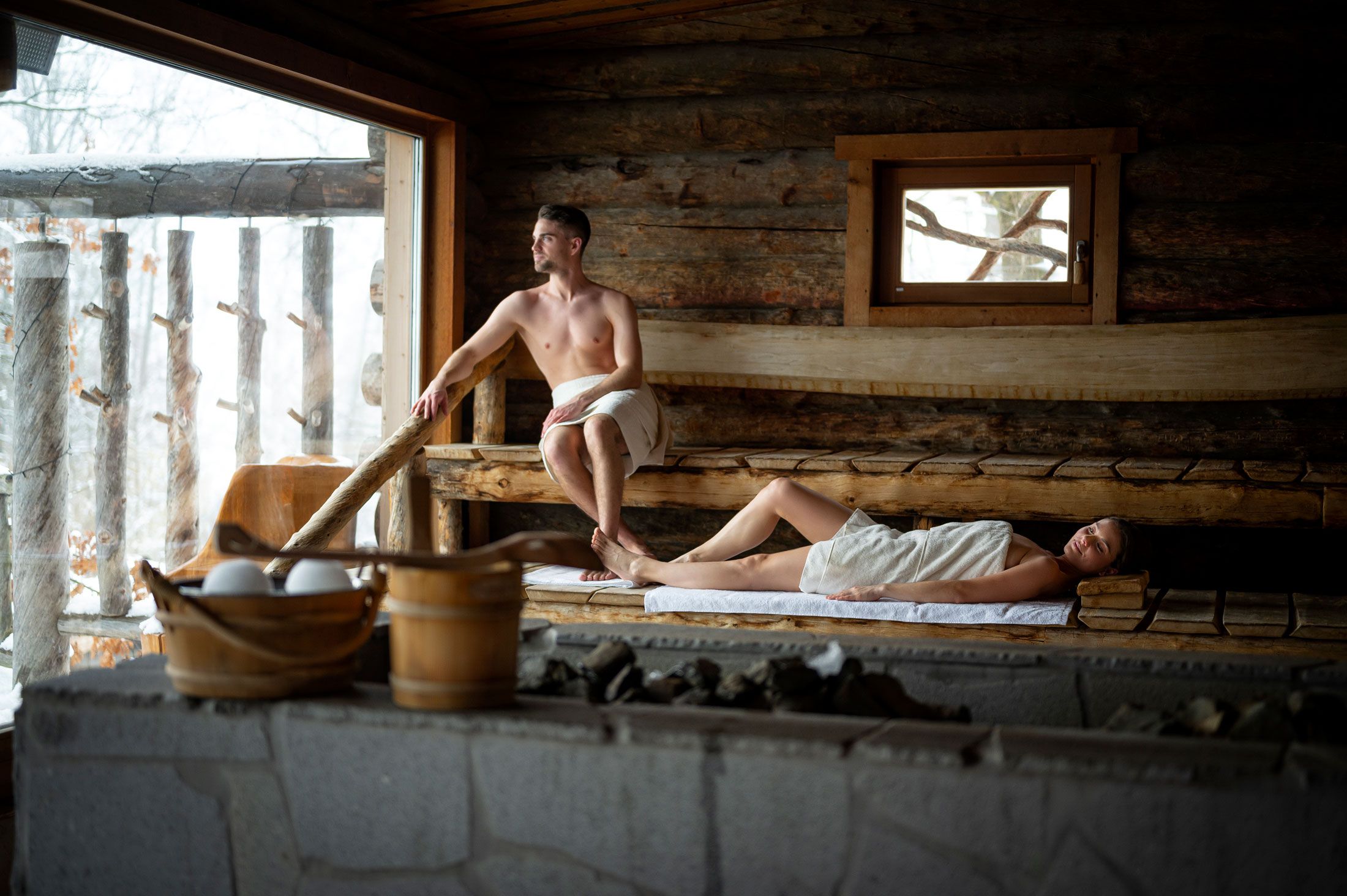 Zwei Personen in einer Holzsauna, eine sitzt, die andere liegt, beide in Handtücher gehüllt.