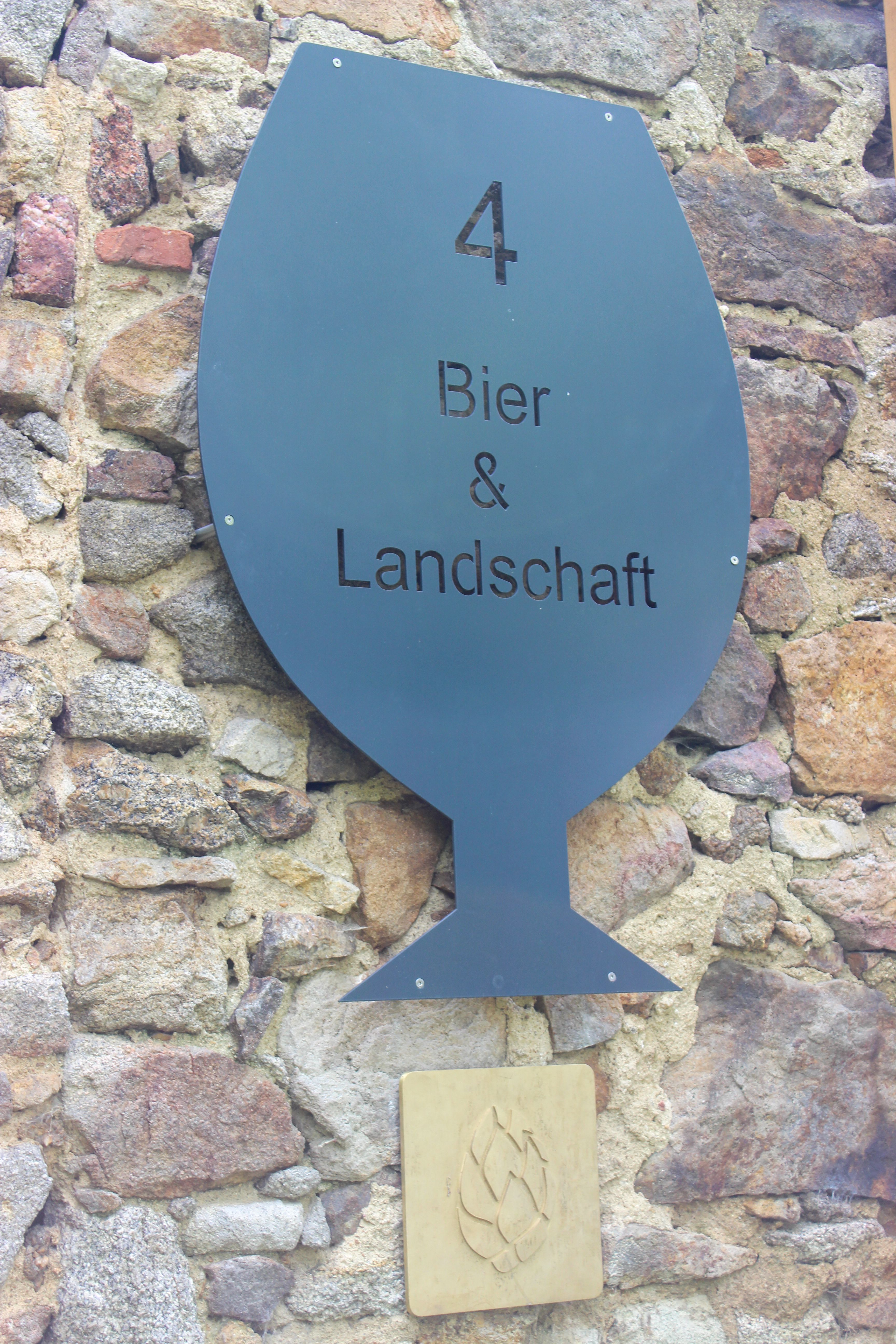 Ein Schild auf einer alten Mauer. Auf dem Schild steht Bier & Landschaft, darunter eine goldene Hpfendolde.