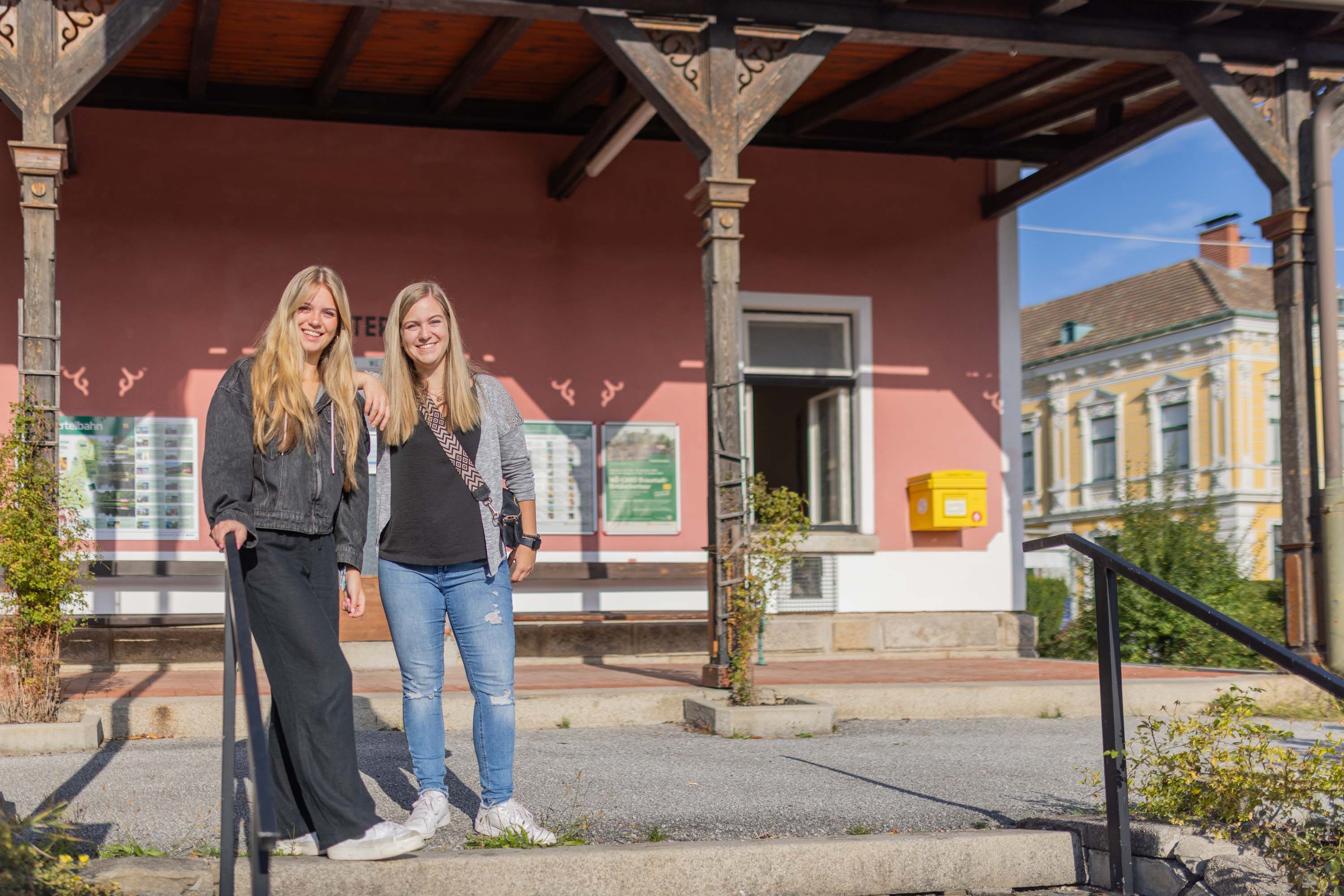 Zwei junge Frauen stehen vor dem Bahnhof von Weitra, der im Hintergrund zu sehen ist.