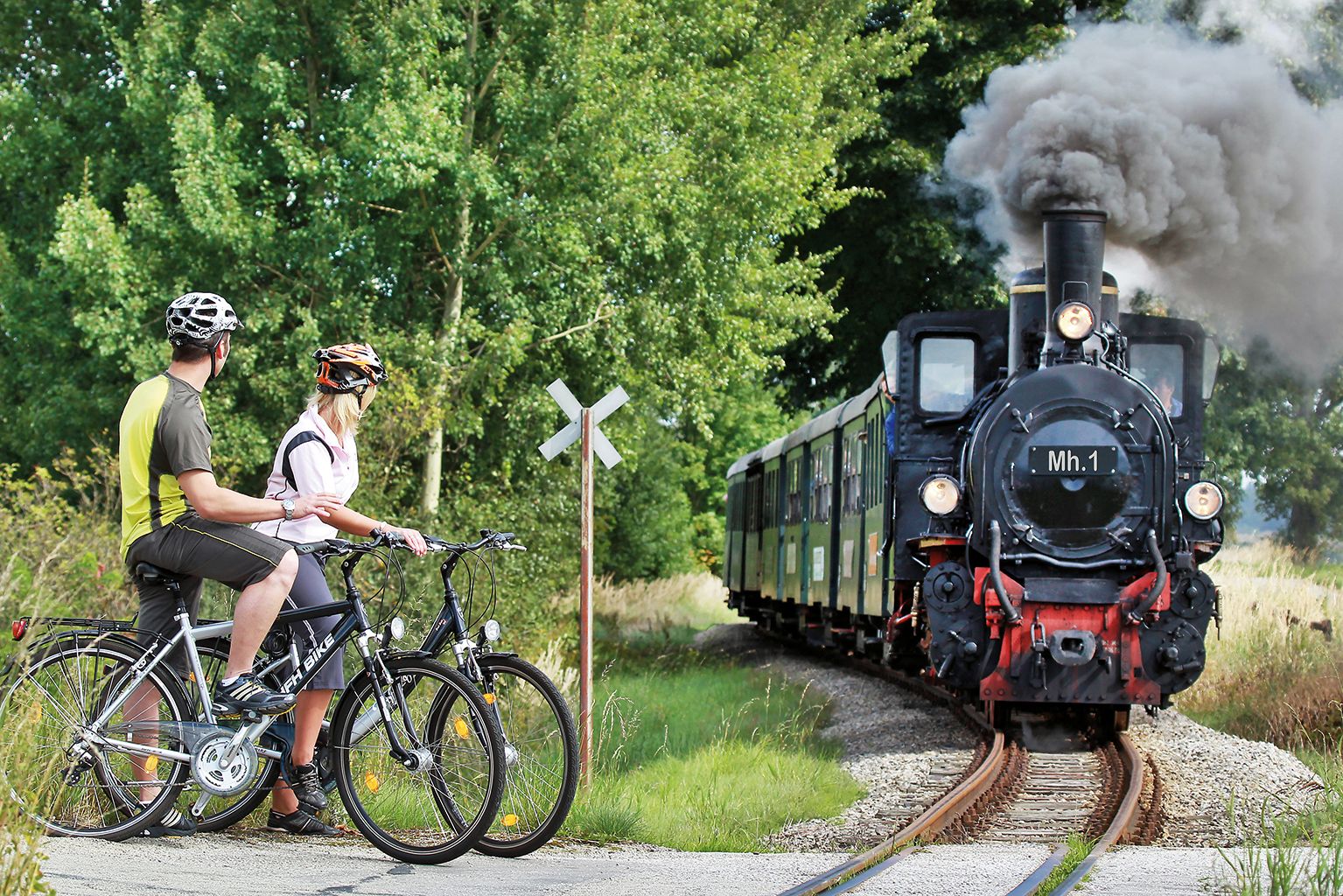 Zwei Radfahrer warten an einem Bahnübergang, während eine Dampflok vorbeifährt.